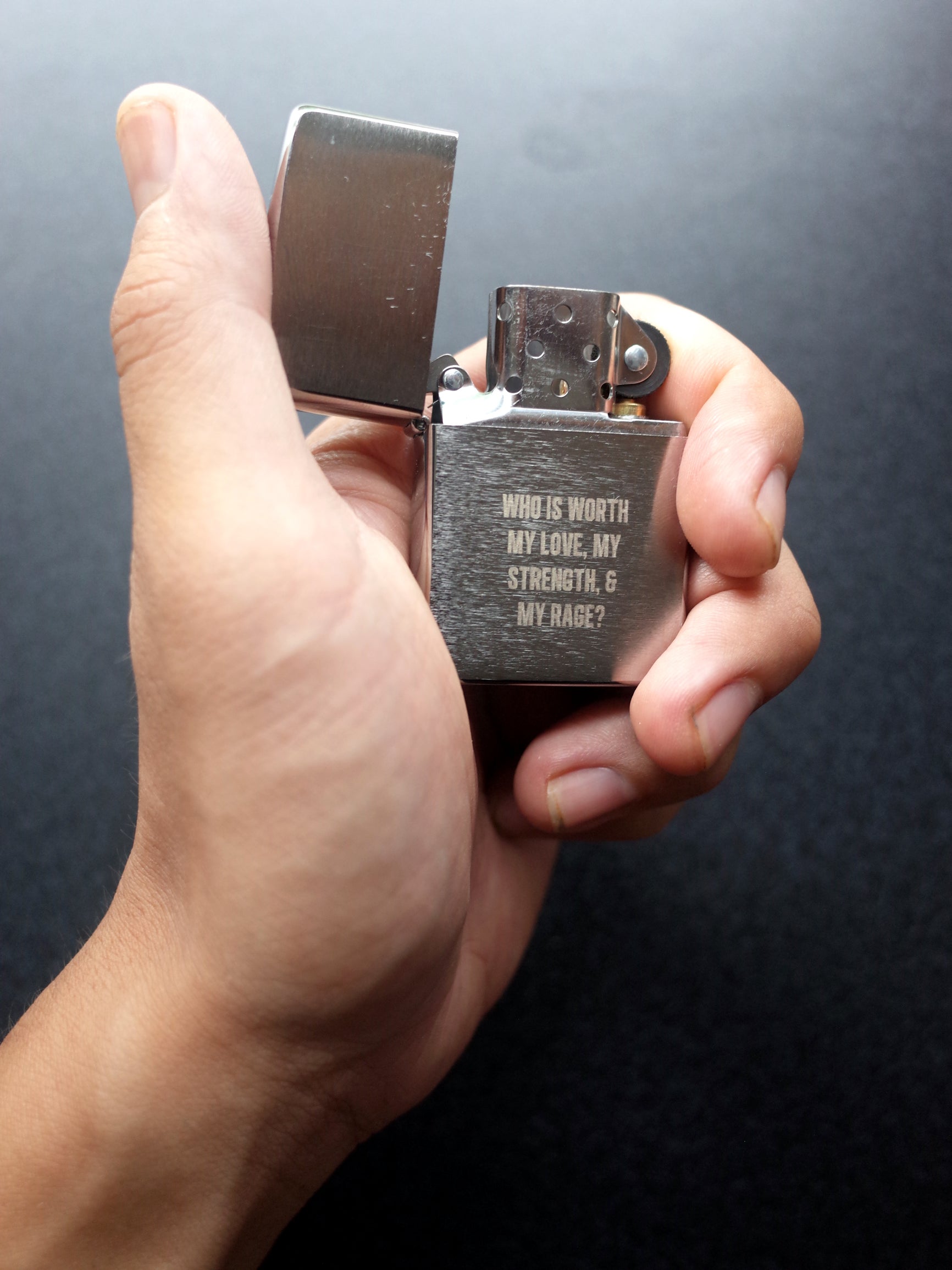 A hand holding an open Zippo style lighter with the engraving "WHO IS WORTH MY LOVE, MY STRENGTH, & MY RAGE?"