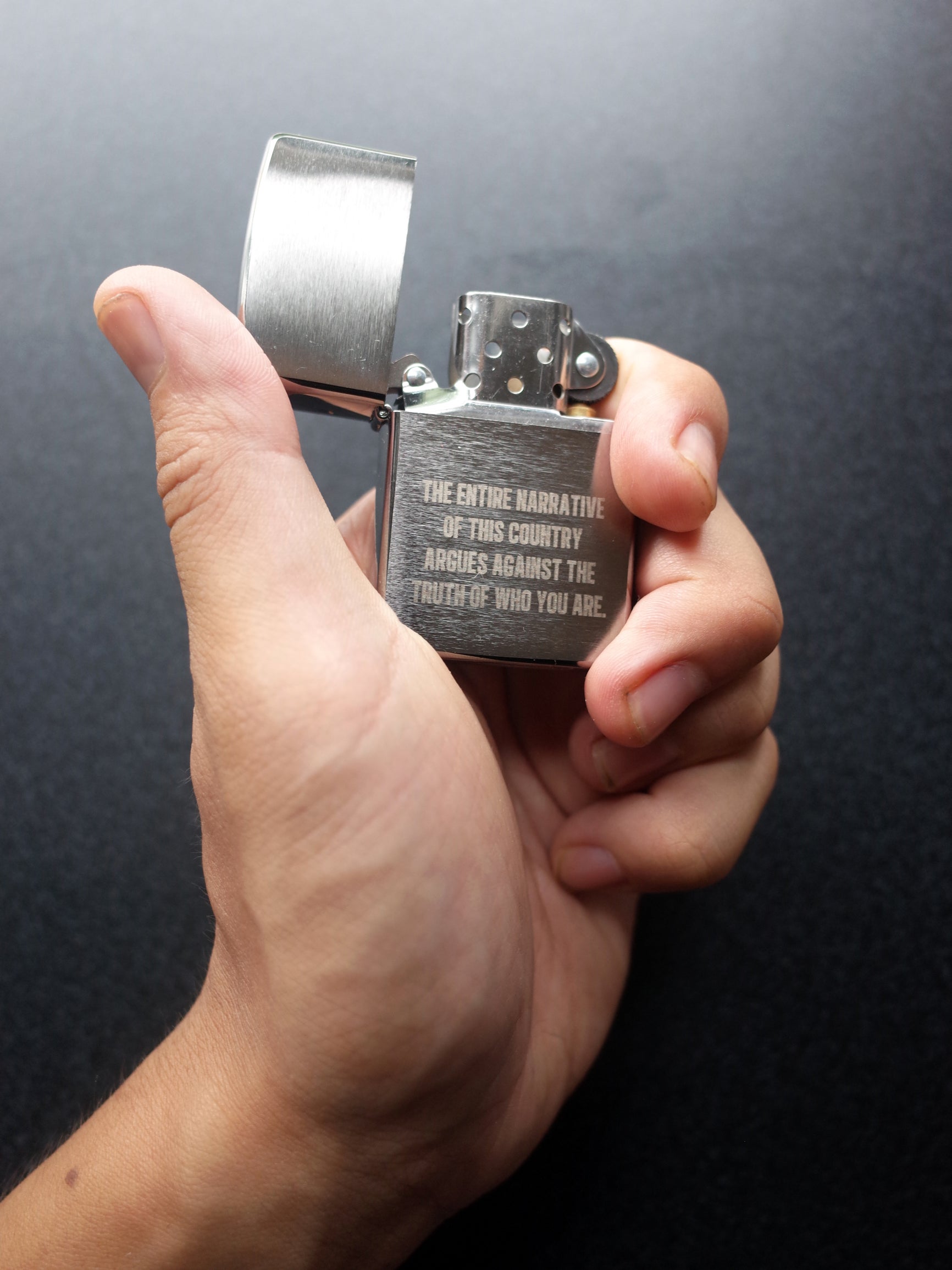 A hand holding an open Zippo style lighter with the engraving "THE ENTIRE NARRATIVE OF THIS COUNTRY ARGUES AGAINST THE TRUTH OF WHO YOU ARE."