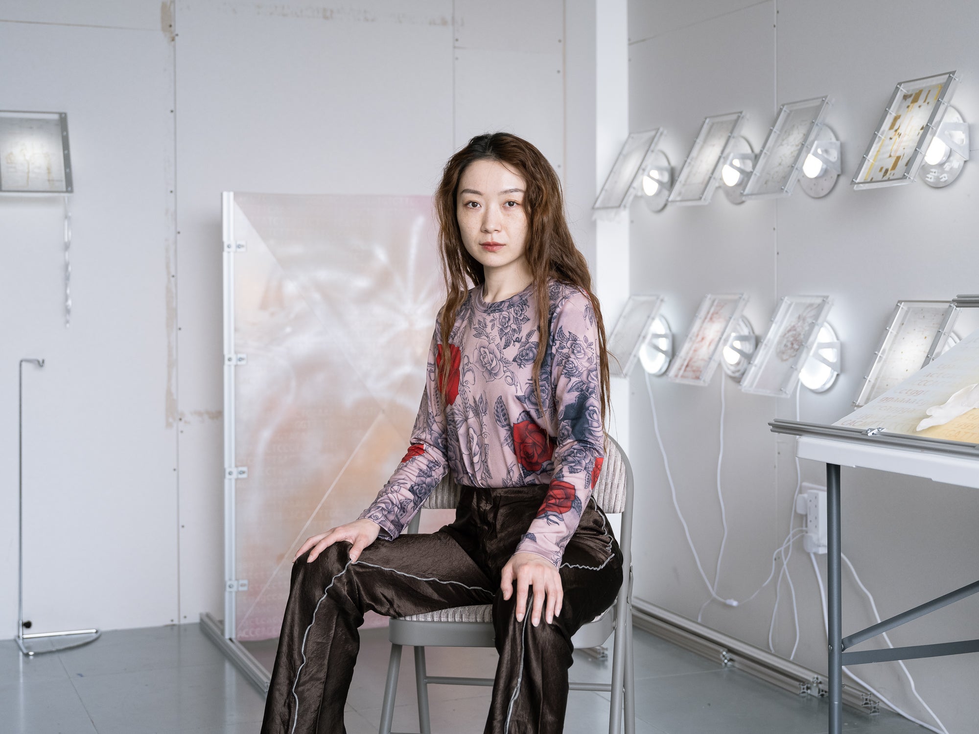 Xin Liu poses sitting on a folding chair in the middle of an art exhibit